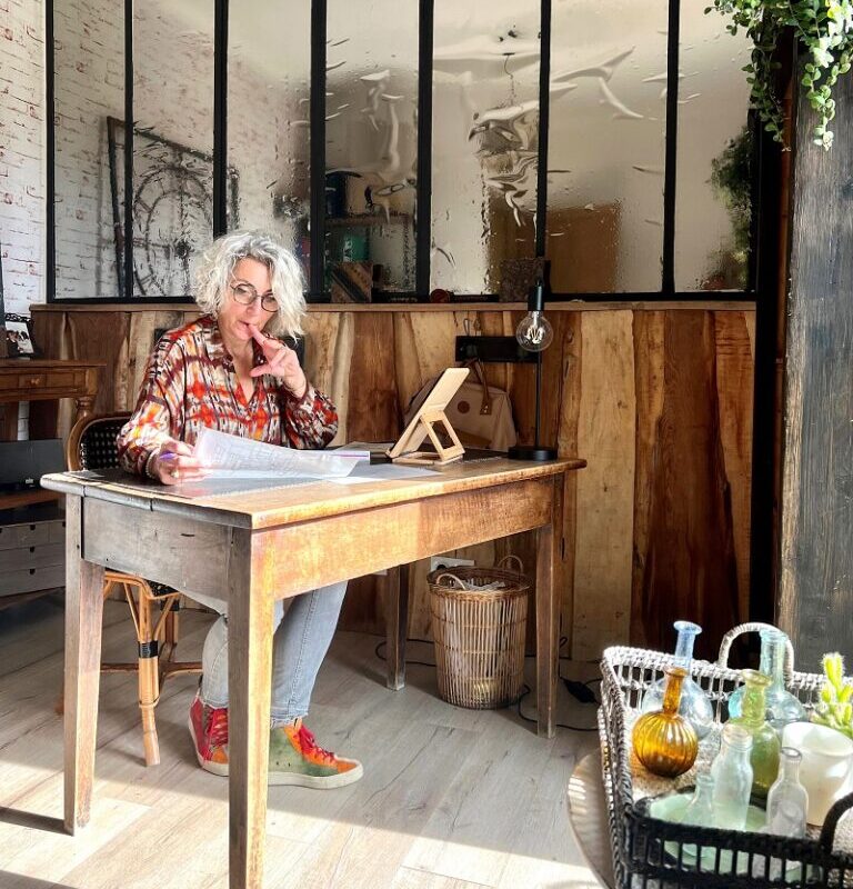 Femme aux cheveux gris en lunettes, concentrée sur des papiers à son bureau en bois. Elle réfléchit, le doigt à la bouche, dans un intérieur chaleureux avec verrière.