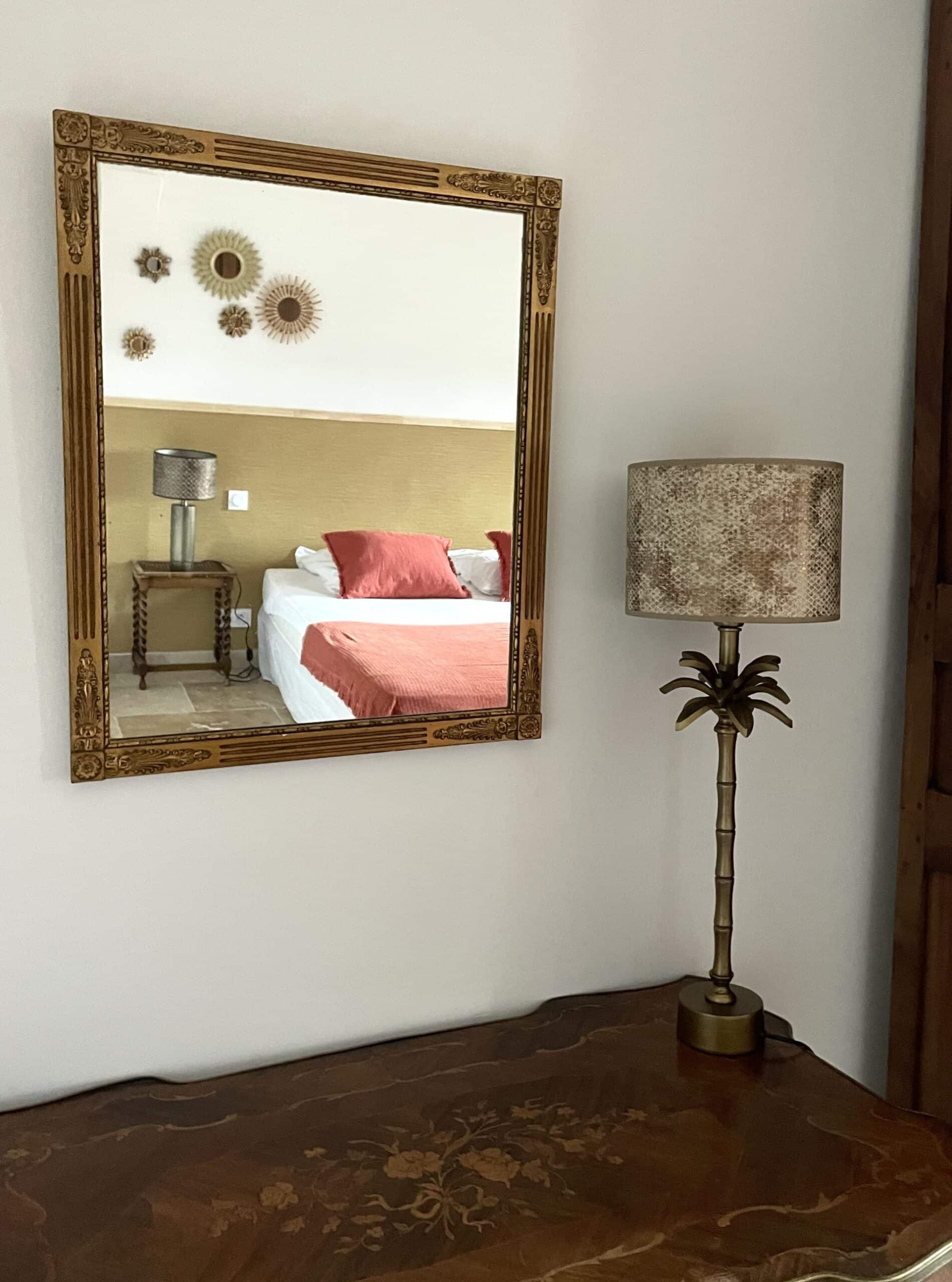 Miroir doré reflétant un lit et lampe palmier exotique sur une commode ancienne marquetée.