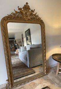 Miroir ancien à cadre doré sculpté reflétant un salon moderne avec canapé gris et tapis en jute.