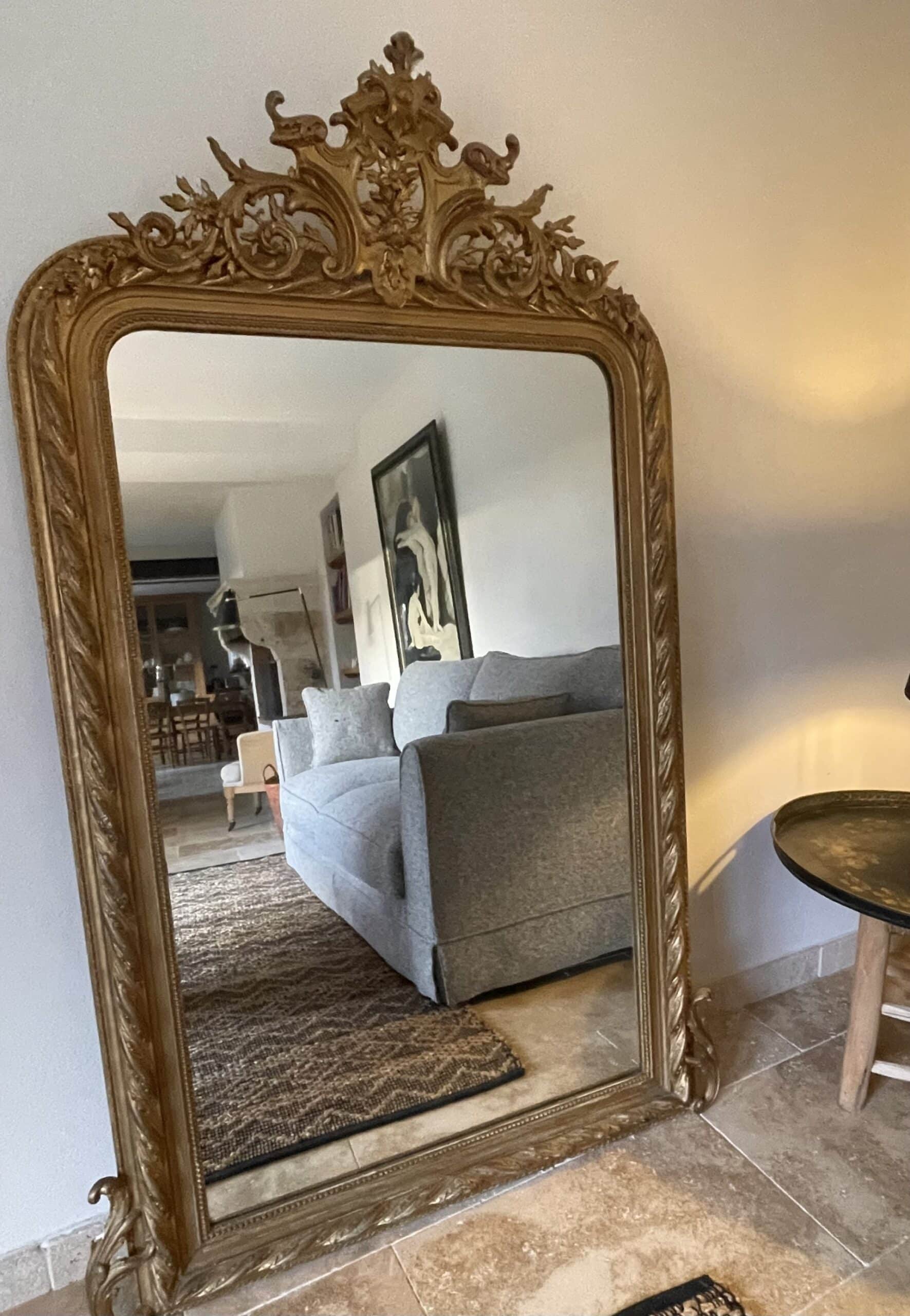 Miroir ancien à cadre doré sculpté reflétant un salon moderne avec canapé gris et tapis en jute.